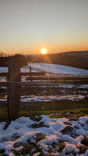 Sonnenuntergang am Horizont, im Vordergrund ist ein Holzzaun zu sehen, dahinter fällt eine verschneite Weide ins Tal hinab. Fotografin: Dorothee