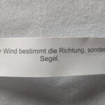 Ein Zettel liegt auf einer weißen Tischdecke. Auf dem Zettel steht der Spruch: Nicht der Wind bestimmt die Richtung, sondern das Segel.