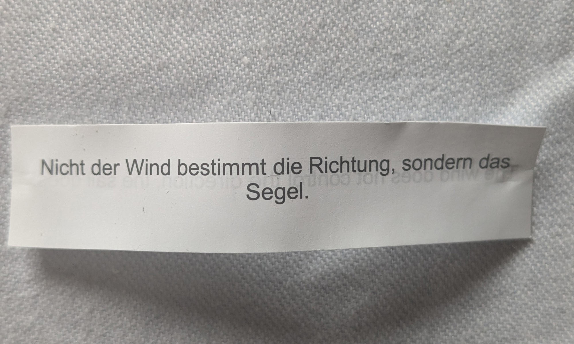 Ein Zettel liegt auf einer weißen Tischdecke. Auf dem Zettel steht der Spruch: Nicht der Wind bestimmt die Richtung, sondern das Segel.