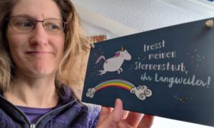 Auf dem Bild ist Dorothee zu sehen, wie sie ein Schild in der Hand hält. Darauf ist ein Einhorn auf einem Regenbogen abgebildet und daneben steht der Spruch: Fresst meinen Sternenstaub, ihr Langweiler