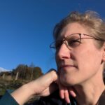 Dorothee sitzt mit dem Kopf auf die Hand gestützt vor blauem Himmel und Wald im Hintergrund