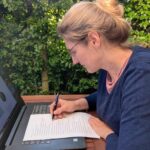 Dorothee sitzt mit Laptop und Papier-To-Want-Liste am Tisch auf der Terrasse