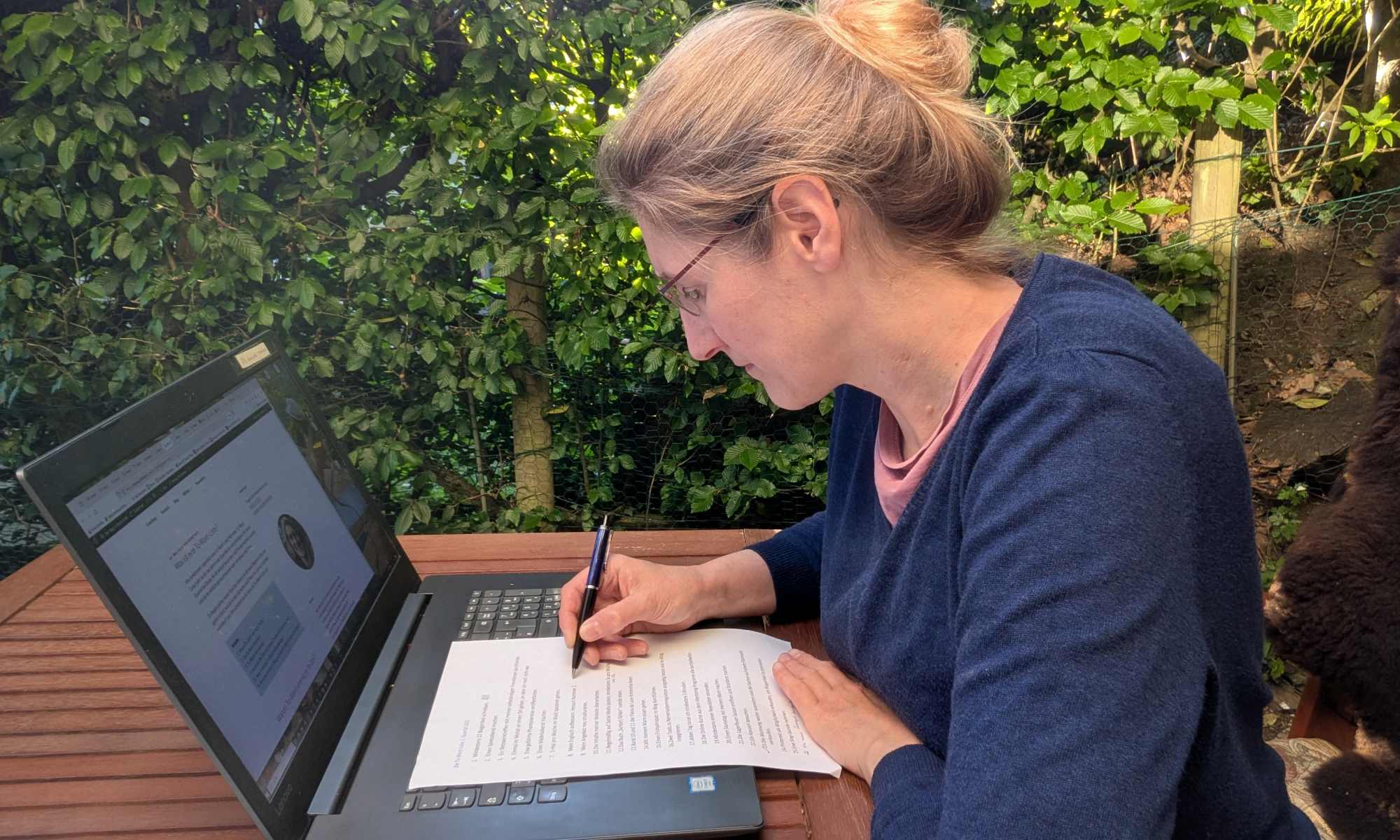 Dorothee sitzt mit Laptop und Papier-To-Want-Liste am Tisch auf der Terrasse