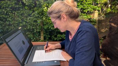 Dorothee auf der Terrasse am Tisch sitzend und schreibend mit Zettel und Stift