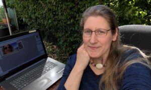 Sichtbar werden mit Dorothee; hier sitzt sie auf der Terrasse mit ihrem Laptop am Tisch und lächelt in die Kamera